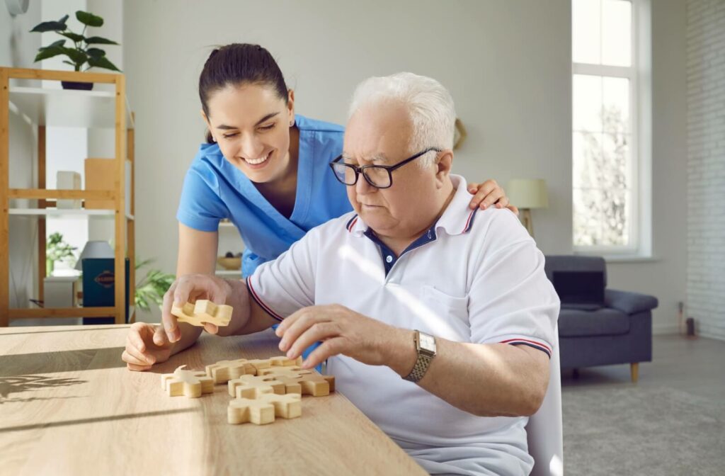 An older man with glasses sitting at a table and doing a jigsaw puzzle with the help of a caregiver