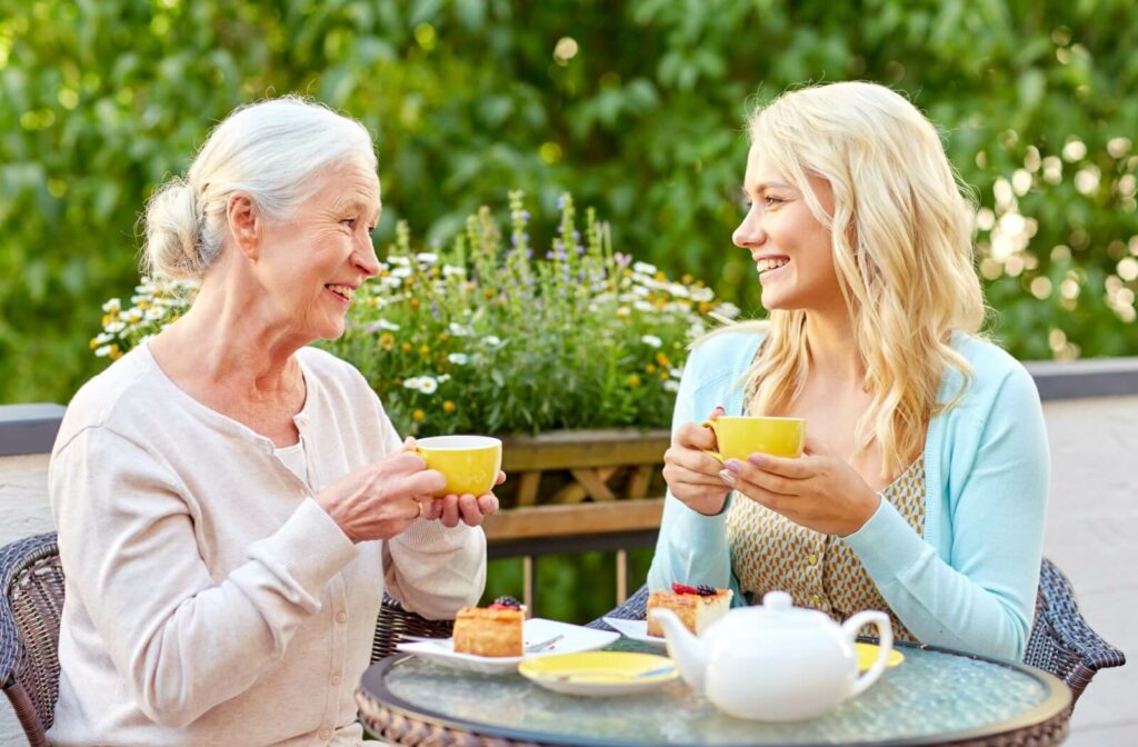 An older woman and her young adult daughter sitting at a table outside holding tea cups and smiling at each other