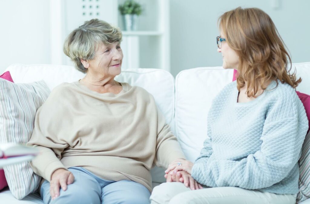 An older woman and her younger adult daughter sitting on a couch holding hands while smiling at each other