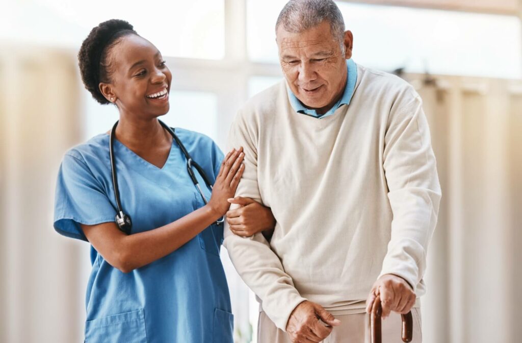 A young female nurse smiling at an older man with a cane and holding his arm while they walk