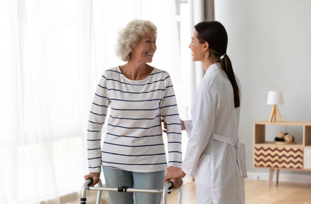 An adult female caretaker and an older woman smiling at each other while they stand in a room at a senior living community.