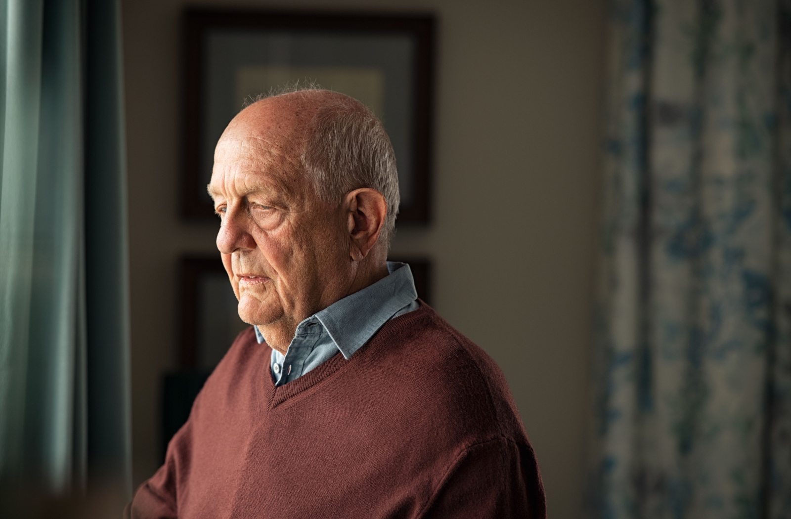 An older adult man looking outside the window with a serious expression.