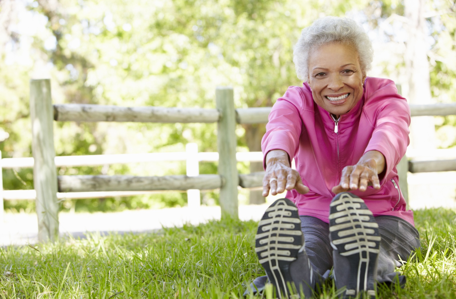 A senior wearing pink and grey athletic wear sits in the grass and stretches their hands to their toes, smiling in the summer sunshine.