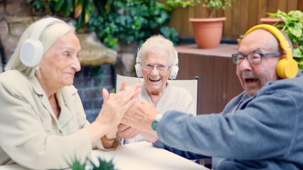 Three older adults laugh and clasp hands while listening to music on headphones.