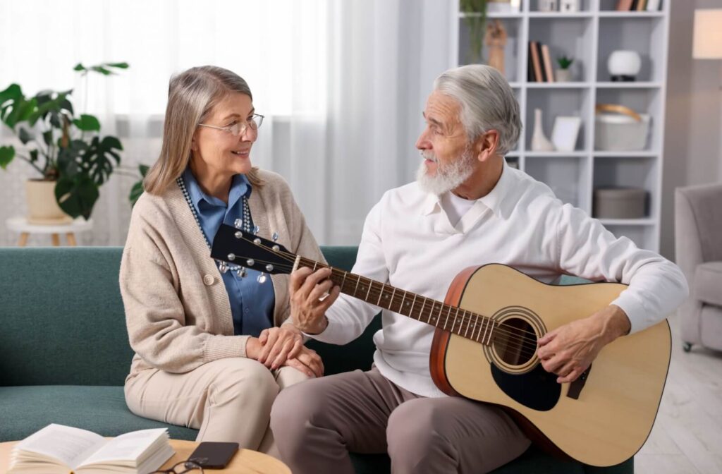 Older adult strumming guitar while sitting with partner in a warm memory care setting focused on dementia support through music